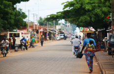 Outdoor street scene with trees, pedestrians, and vehicles lining the sandy road